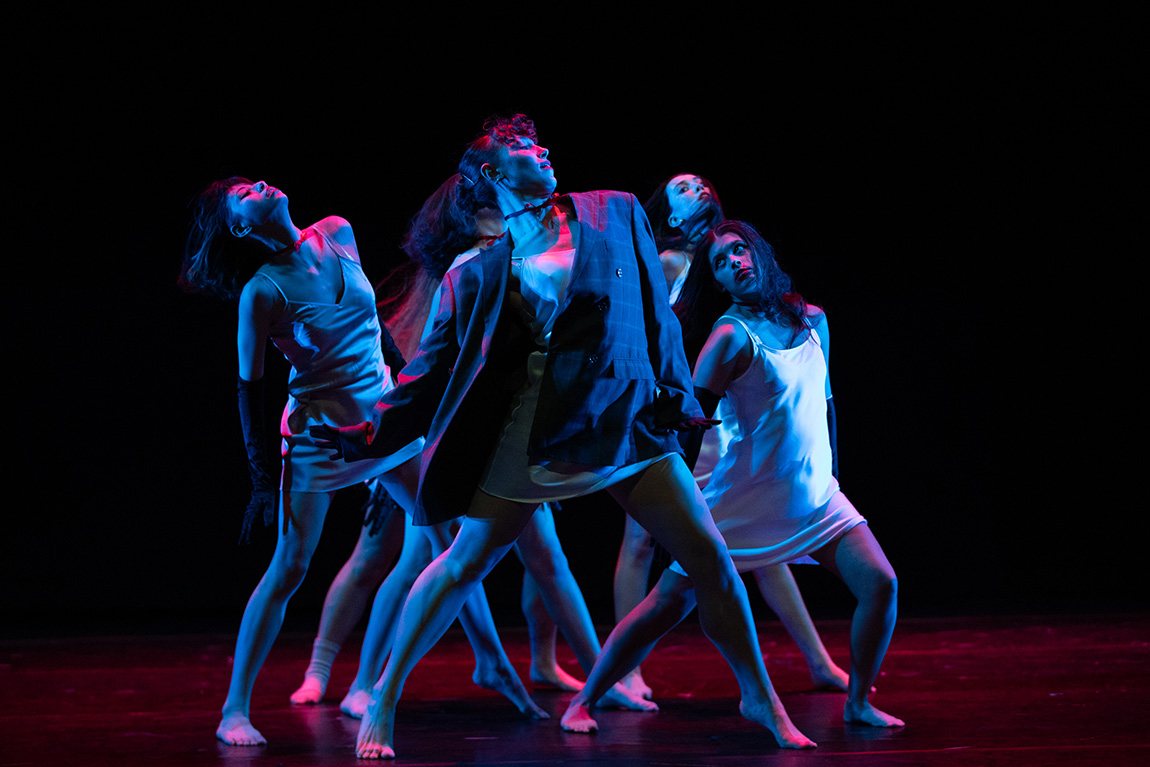 Production photo from a dance performance, featuring six dancers in costume, under stage light. Photo by Marco Calderon.